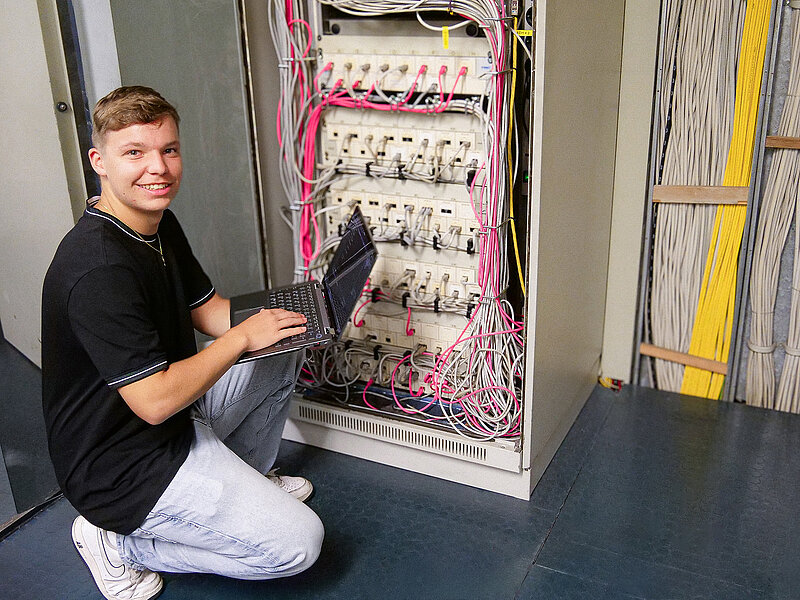 Fachinformatiker-Azubi bei RUD arbeitet mit Laptop an einem Serverschrank mit Kabeln.