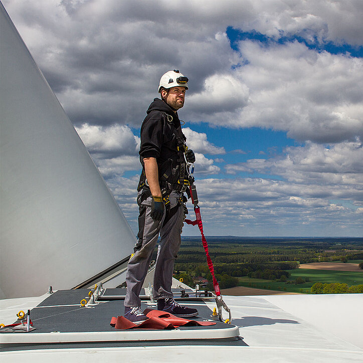 Ein Techniker mit Sicherheitsausrüstung steht auf einer Windkraftanlage und ist über ein rotes Verbindungsmittel mit einem PSA-Anschlagpunkt gesichert. Im Hintergrund sind Wälder und Felder unter einem wolkigen Himmel zu sehen.