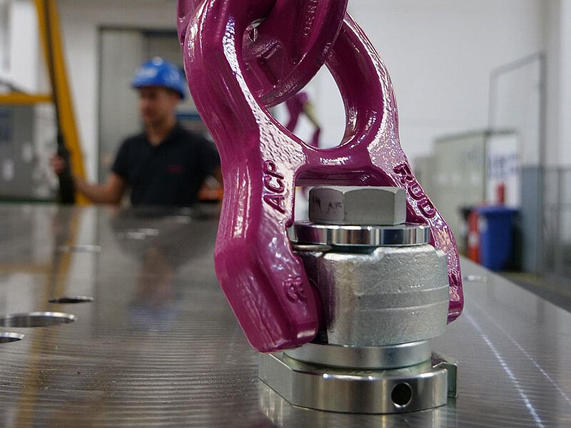 Montierter RUD-Anschlagpunkt bei Hebevorgang auf Metallfläche, mit Arbeiter im Hintergrund.