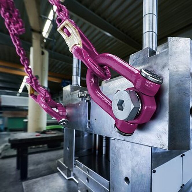 Ein metallischer Maschinenbauteil wird mithilfe eines pinken RUD ACP-TURNADO Anschlagpunkts und einer angeschlossenen Kette gehoben oder gesichert. Die glänzende Oberfläche der massiven Komponente sowie die präzise Verschraubung mit der Sechskantschraube verdeutlichen die hohe Belastbarkeit und Sicherheitsanforderung der Anwendung. Im Hintergrund ist eine industrielle Werkhalle zu erkennen.