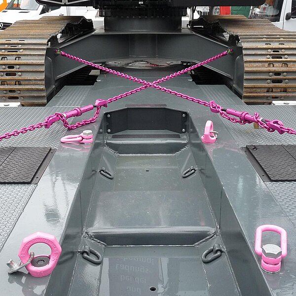 Eine schwere Baumaschine ist auf einem Tieflader mit pinkfarbenen RUD-Zurrketten und Anschlagpunkten gesichert. Die Ketten sind über Kreuz gespannt und fixieren das Fahrzeug sicher auf der Ladefläche.
