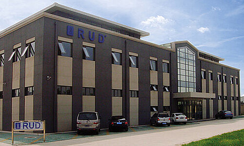 RUD Lifting Beijing Co. Ltd, modernes mehrstöckiges Firmengebäude mit Glasfassade am Eingang und RUD Logo, mehrere Autos auf dem Parkplatz.
