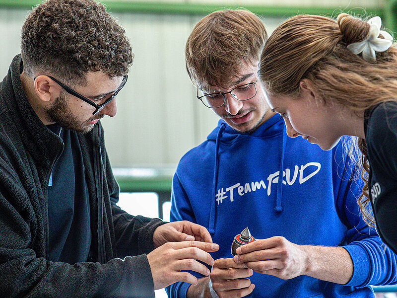 Drei RUD Werkstudenten betrachten gemeinsam ein metallisches Bauteil und besprechen dessen Details.