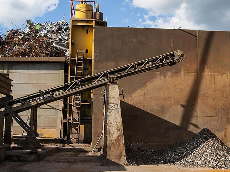 Ein Förderband transportiert zerkleinertes Metall vor eine Recyclinganlage, im Hintergrund ist ein großer Schrottberg zu sehen.