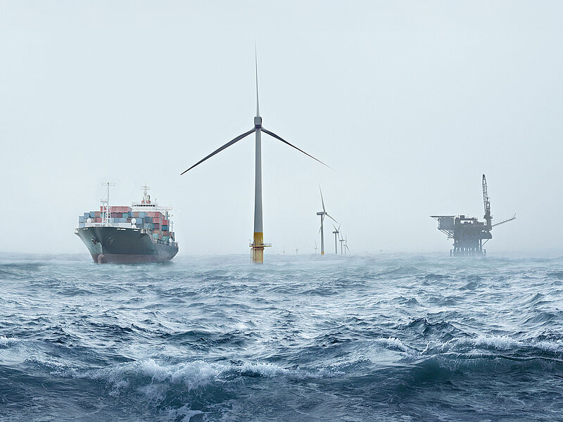 Container-Schiff auf dem Meer, umgeben von Offshore-Windkraftanlagen und einer Ölplattform im Hintergrund.