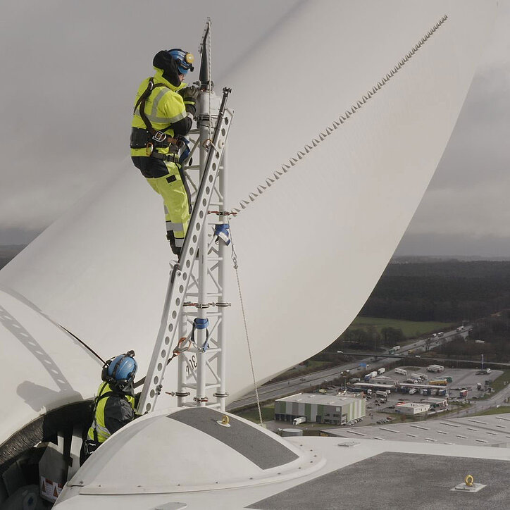 Zwei Fachkräfte in Schutzkleidung und mit PSA gesichert arbeiten auf der Gondel einer Windkraftanlage. Einer der beiden steht auf einer temporär befestigten Arbeitsplattform an einem Rotorblatt. Im Hintergrund sind Felder, Wälder und ein Industriegebiet mit Parkplätzen zu sehen.