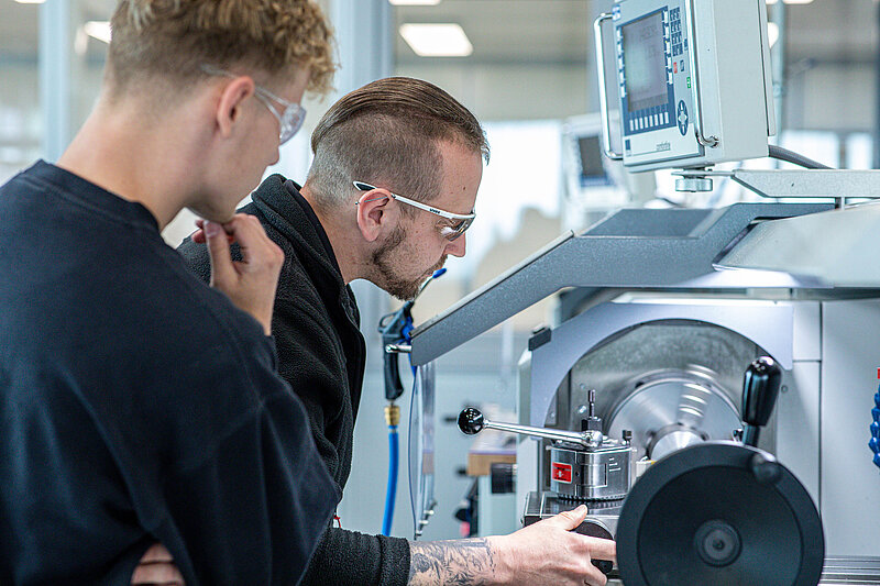 In einer modernen Werkhalle arbeitet ein Rud Mitarbeiter mit Schutzbrille an einer CNC-Maschine, während ein weiterer RUD Mitarbeiter mit Schutzbrille ihm aufmerksam dabei zusieht.