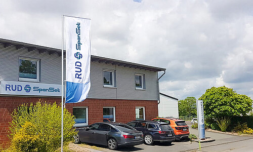 Außenansicht der RUD SpanSet GmbH Soest mit Firmenflagge, Logo und geparkten Autos vor dem Gebäude.