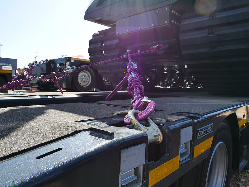 Ein schweres Fahrzeug mit Ketten von RUD auf einem Tieflader der Marke Goldhofer gesichert; im Vordergrund sind pinke Kettenglieder und ein Zurrhaken in einer Verankerung sichtbar.
