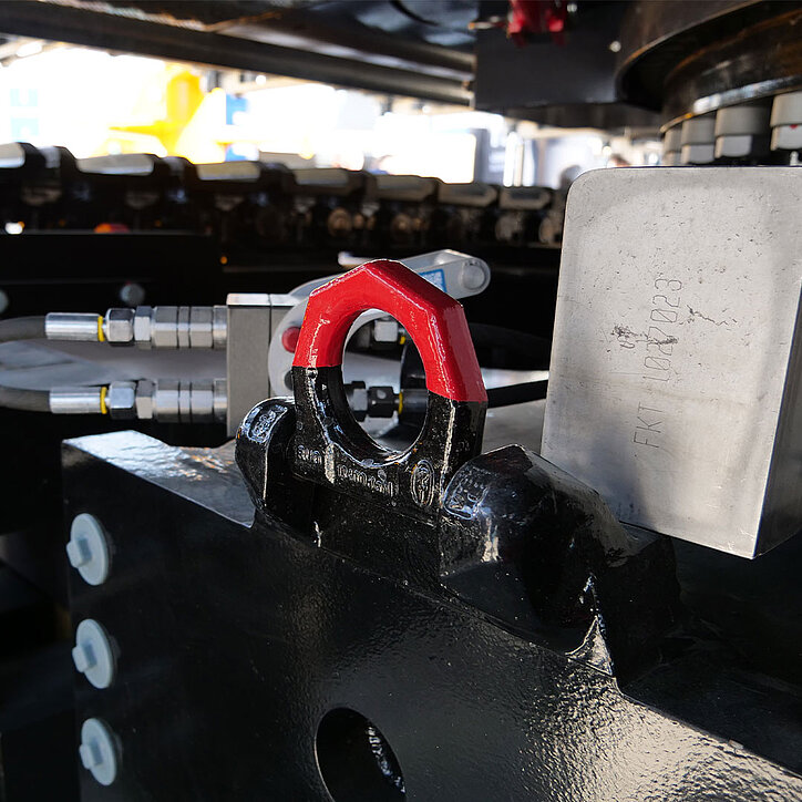 Schwarz-rot lackierter Zurrpunkt von RUD, fest verschweißt an einer robusten Maschinenstruktur mit hydraulischen Anschlüssen im Hintergrund. Der Zurrpunkt befindet sich in einem industriellen Umfeld unterhalb eines Kettenfahrzeugs.