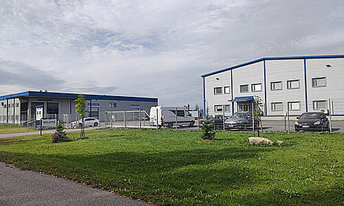 Außenansicht des Erlatek Oy Standorts mit modernen Hallengebäuden und Parkplatz.