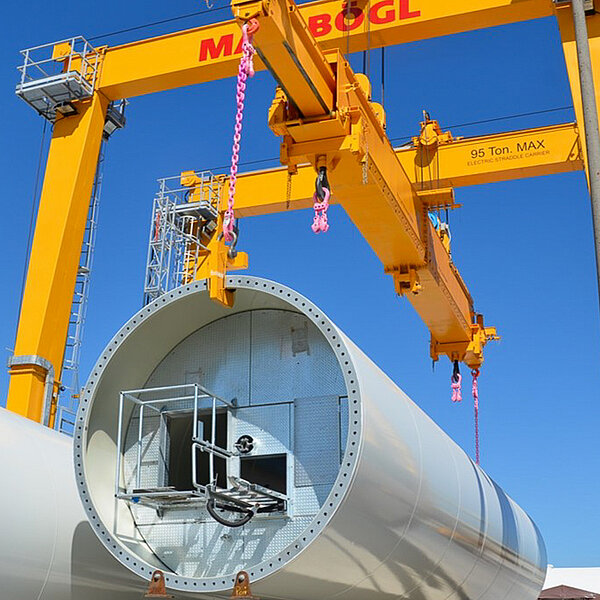 Ein 95-Tonnen-Portalkran hebt mit RUD Anschlagpunkten und -ketten ein Turmsegment für eine Windkraftanlage an.