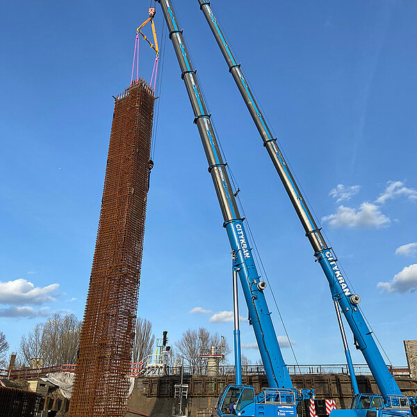 Zwei CITYKRAN-Fahrzeuge heben einen hohen Gitterkorb mithilfe von RUD Anschlagmitteln auf einer Baustelle in Berlin.
