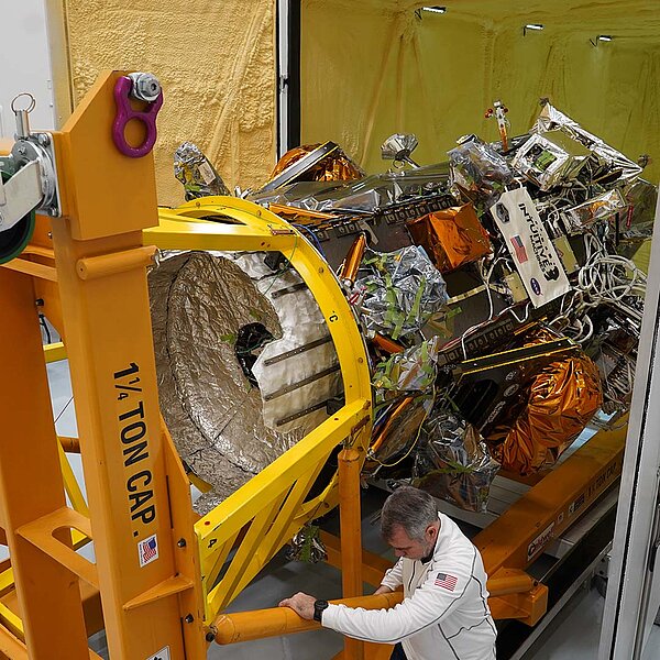 Ein NASA-Mitarbeiter montiert ein mit goldener Isolierfolie umhülltes Raumfahrtmodul, gesichert mit einem violetten RUD-Anschlagpunkt.