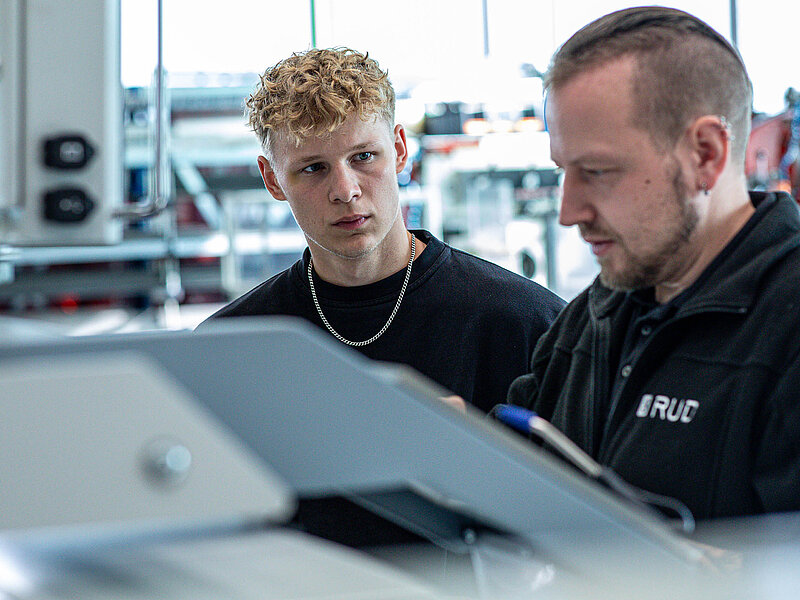 Ein junger Mann beobachtet aufmerksam einen Ausbilder bei der Arbeit an einer Maschine in einer Werkhalle.