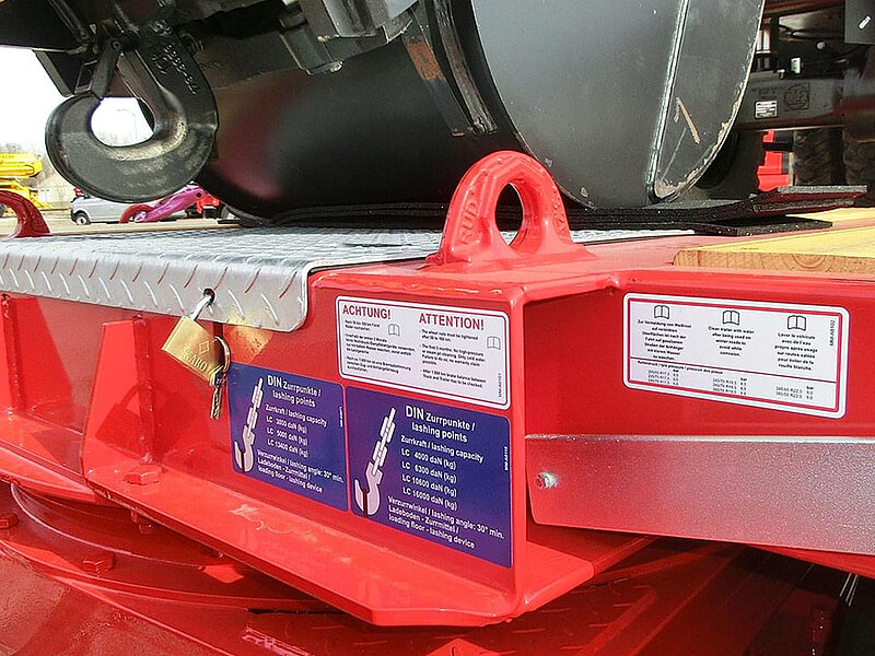 Zurrpunkt mit Lastangaben an rotem Anhänger, inklusive Sicherheitshinweisen und DIN-Angaben.