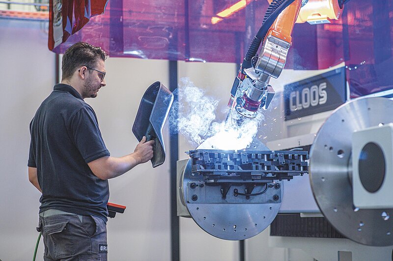 Ein Mitarbeiter beobachtet mit Schweißerschutzschild in der Hand, wie ein Schweißroboter in einer Werkhalle unter Funkenflug und Rauch ein Metallteil automatisiert verschweißt.