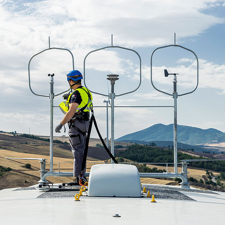 Ein Techniker mit Schutzhelm und Sicherheitsausrüstung steht auf dem Dach einer Windkraftanlage und ist mit einem PSA-Anschlagpunkt gesichert. Im Hintergrund sind Hügel und ein teilweise bewölkter Himmel zu sehen.