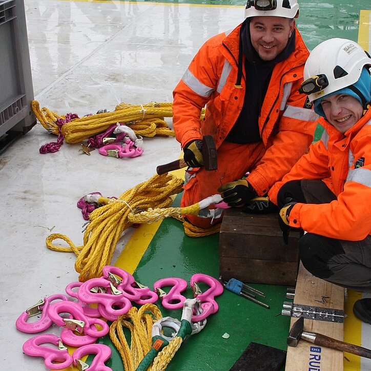 Zwei Monteure montieren pinke RUD-Hebepunkte an gelbe Anschlagseile auf einem Offshore-Schiff.