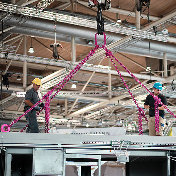 Arbeiter sichern ein schweres Bauteil mit pinken RUD-Hebeketten an einem Hallenkran.