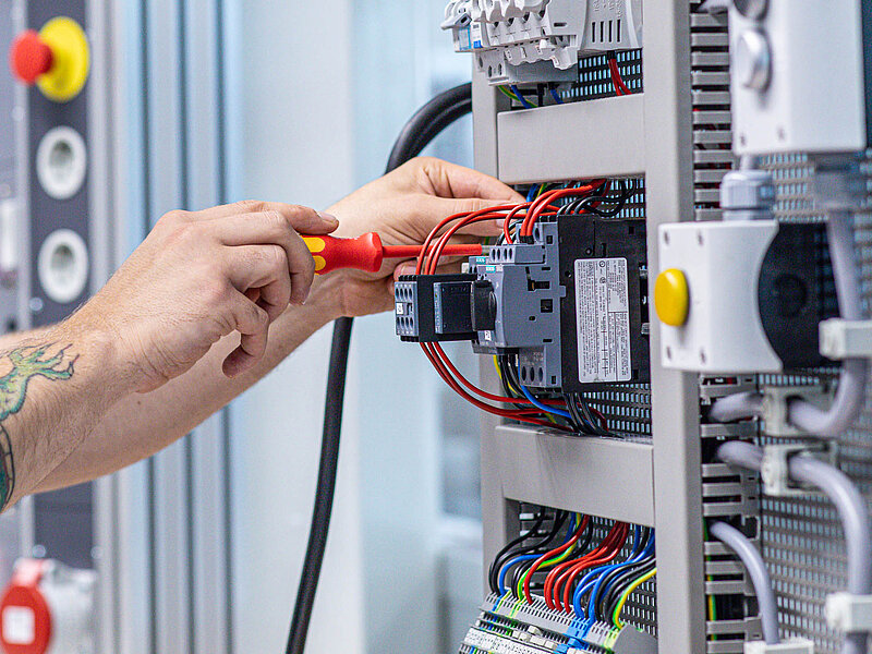 Ein Elektroniker arbeitet mit Schraubenzieher und Kabeln an einem offenen Schaltschrank mit elektrischen Bauteilen.