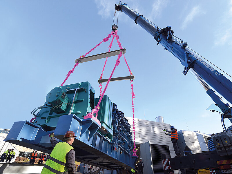 Großes Maschinenmodul wird mit einem Kran und pinkfarbenen Ketten gehoben, während Arbeiter den Vorgang überwachen.