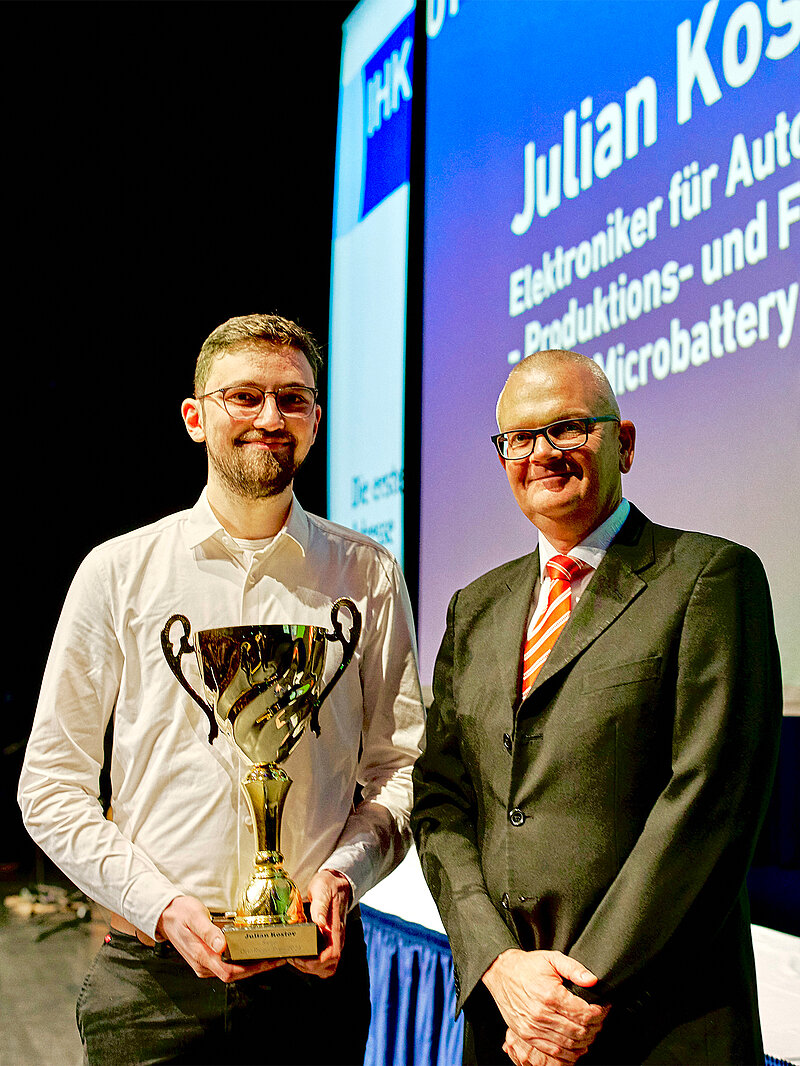 Preisverleihung: Ein Mann in weißem Hemd hält einen goldenen Pokal, neben ihm steht ein älterer Mann im Anzug; im Hintergrund ist eine Leinwand mit dem IHK-Logo und Namen zu sehen.