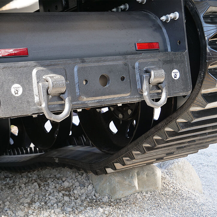 Zwei geschweißte Zurrpunkte von RUD an der Rückseite eines Kettenfahrzeugs, montiert an einer massiven Metallstruktur über dem Laufwerk mit Gummiketten. Die Zurrpunkte sind als stabile Befestigungslösungen sichtbar markiert.