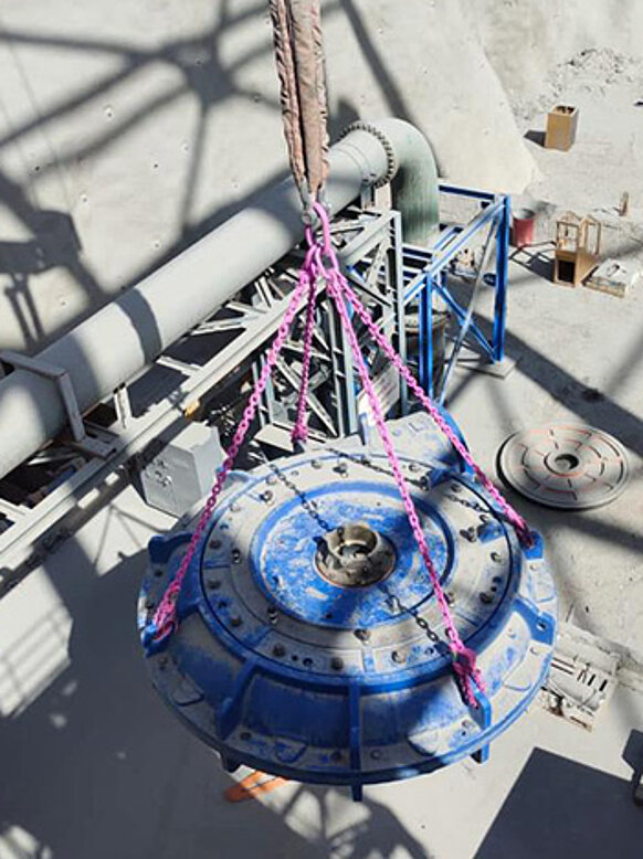 Eine massive, blaue Industriepumpe wird von oben gezeigt, während sie mit vier pinken RUD-Zurrketten angehoben wird. Die Ketten sind an einem zentralen Hebepunkt gebündelt. Im Hintergrund sind industrielle Anlagen, Rohre und Metallkonstruktionen zu sehen. Die Szene spielt in einem staubigen Außenbereich einer Industrieanlage.