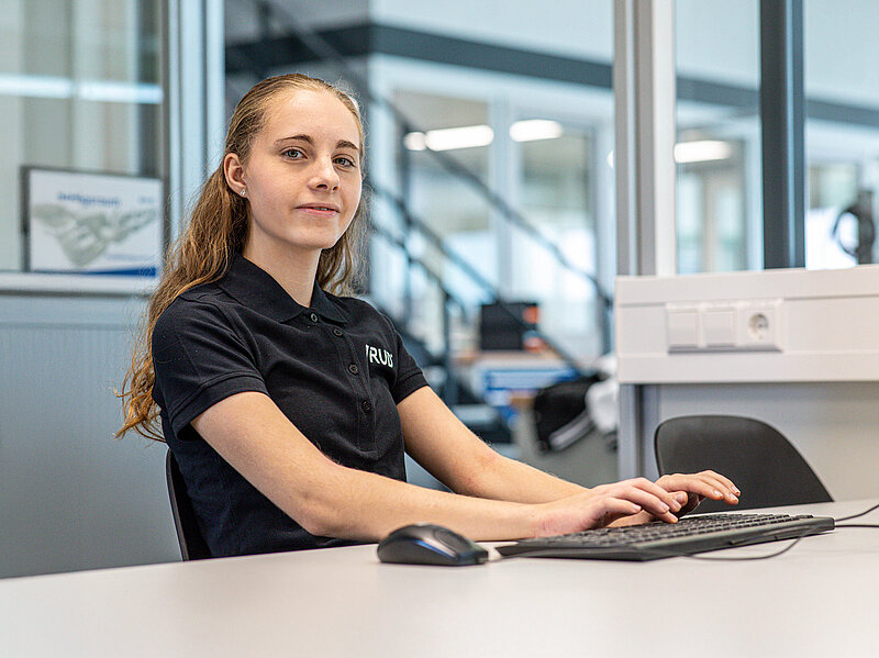 Junge Frau in schwarzem RUD Polohemd sitzt am Schreibtisch und arbeitet an Computer mit Tastatur und Maus in einem modernen Büro.