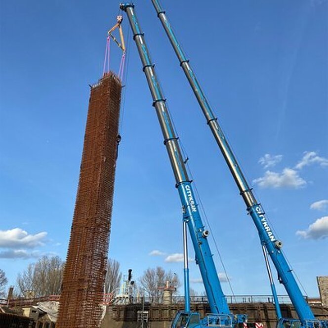 Zwei CITYKRAN-Fahrzeuge heben einen hohen Gitterkorb mithilfe von RUD Anschlagmitteln auf einer Baustelle in Berlin.