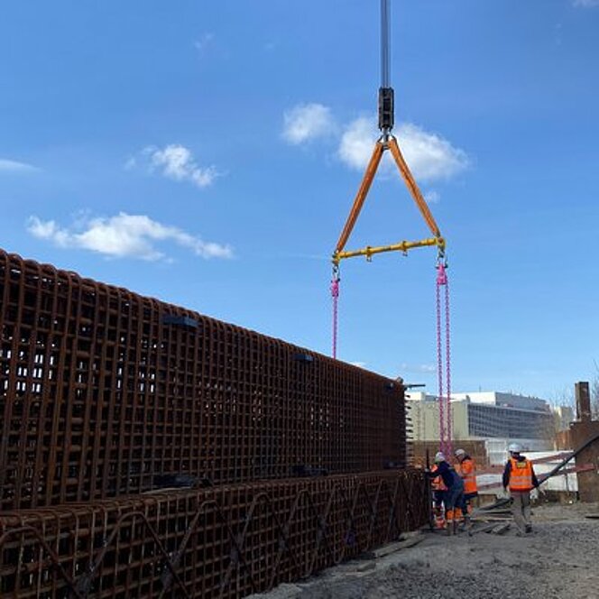 Arbeiter befestigen pinke RUD ChainBlocks mit gelbem Spreizträger an einem großen Gitterkorb auf der Baustelle.