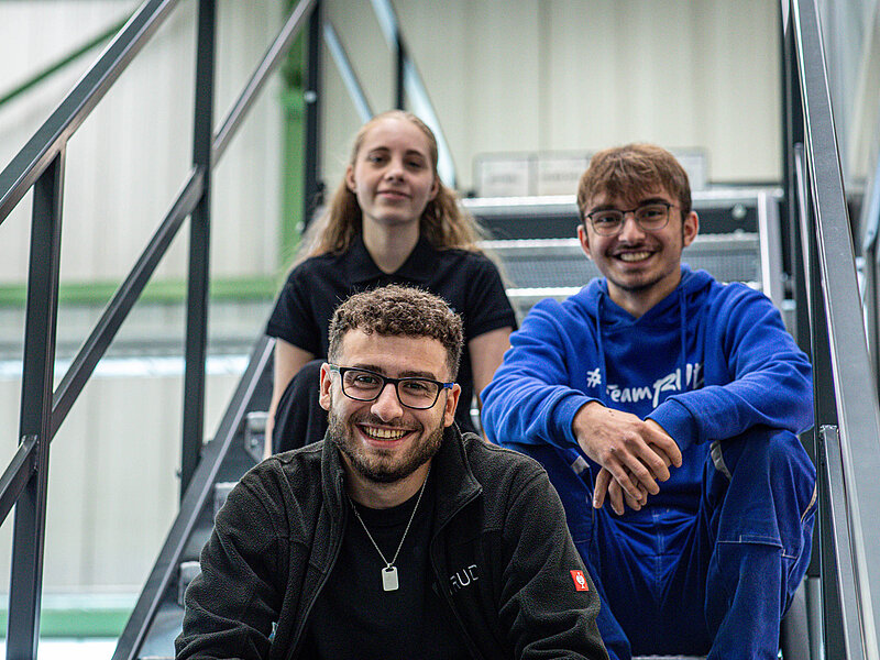 Drei junge RUD Mitarbeitende sitzen lächelnd auf einer Treppe in einem industriellen Arbeitsumfeld.