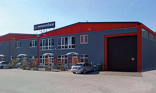 Firmengebäude von Vodušek d.o.o. mit grauer und roter Fassade sowie dem Firmenschild auf dem Dach.