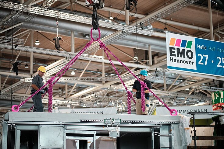 Arbeiter befestigen pinke RUD-Hebeketten an einer Maschine während der EMO Hannover Messe.