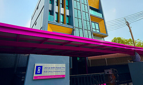 RUD Lifting (Thailand) Co., Ltd., modernes mehrstöckiges Bürogebäude mit gelben Fassadenelementen und auffälligem pinkem Vordach.