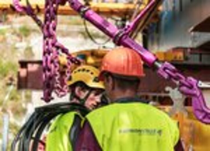 Zwei Bauarbeiter in Sicherheitskleidung überprüfen eine festgezurrte RUD ICE-Zurrkette an einem schweren Stahlbauteil.