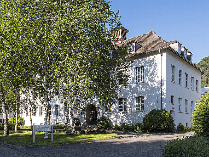 Das weiße RUD Campus-Gebäude mit Satteldach und vielen Fenstern, umgeben von Bäumen und Grünflächen an einem sonnigen Tag.