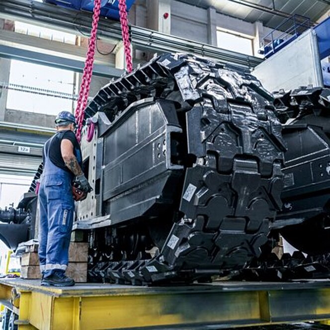 Ein schweres, kettengetriebenes Fahrzeugbauteil wird mithilfe eines pinken RUD ACP-TURNADO Anschlagpunkts und entsprechender Ketten gehoben. Ein Monteur in blauer Arbeitskleidung und Helm überwacht den Vorgang in einer industriellen Fertigungshalle.