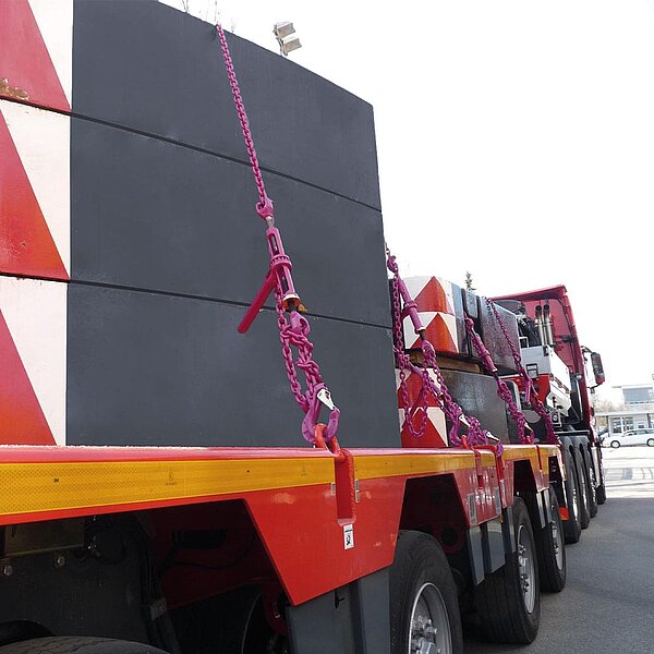 Schwere Lasten werden mit pinkfarbenen Kettenzurrsystemen auf einem roten Tieflader gesichert. Die Ketten sind an massiven Betonblöcken befestigt und über Spannelemente gespannt. Im Hintergrund ist eine rote Schwerlastzugmaschine zu sehen.