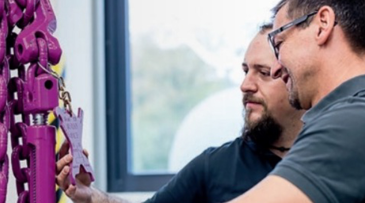 Zwei Fachkräfte prüfen gemeinsam ein RUD-Kettengehänge mit Prüfplakette in heller Arbeitsumgebung.