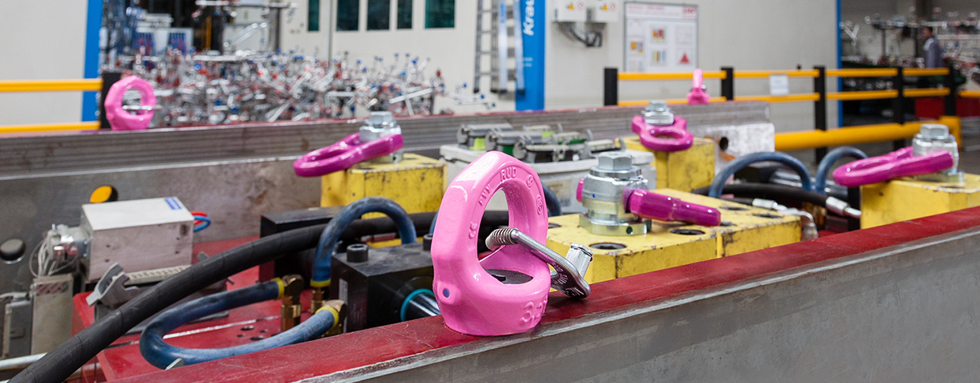 RUD-Anschlagpunkte in Pink montiert auf gelben Vorrichtungen in einer Industriehalle mit Maschinen.