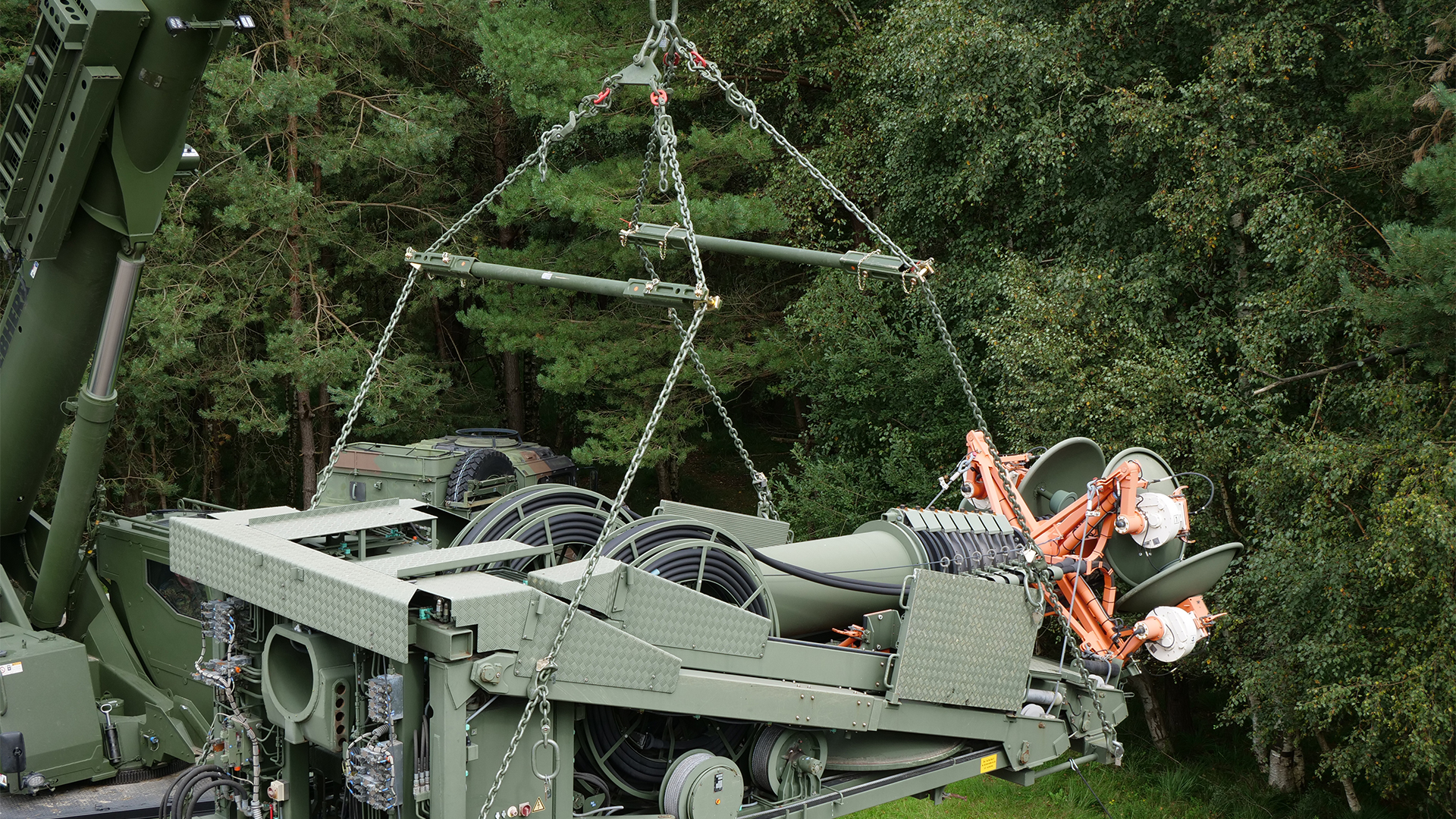 Militärisches Gerät wird mit Kettenhebevorrichtung aus einem Fahrzeug gehoben, umgeben von Waldlandschaft.