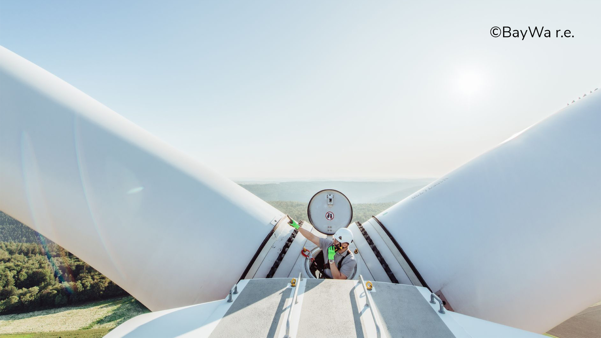 Techniker arbeitet auf der Spitze eines Windkraftwerks, Blick auf die Rotorblätter und die umliegende Landschaft.