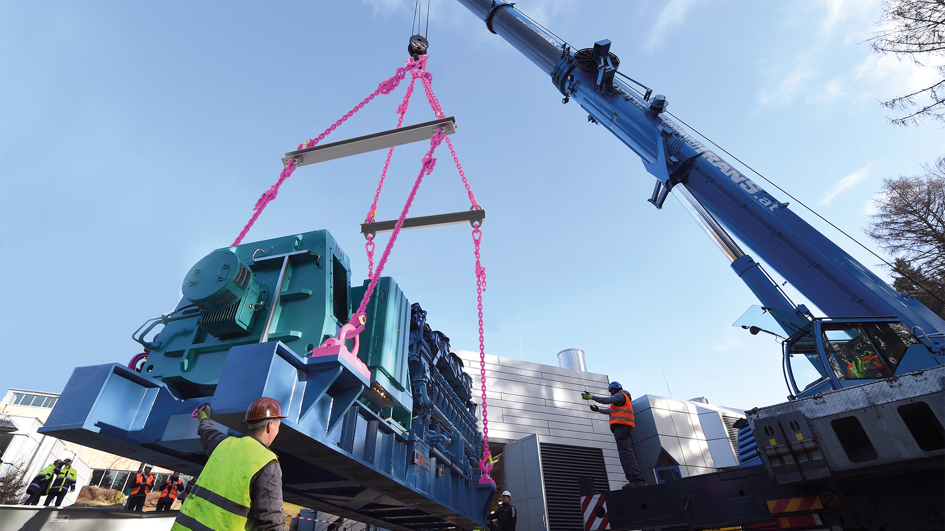 Großes Maschinenmodul wird mit einem Kran und pinkfarbenen Ketten gehoben, während Arbeiter den Vorgang überwachen.