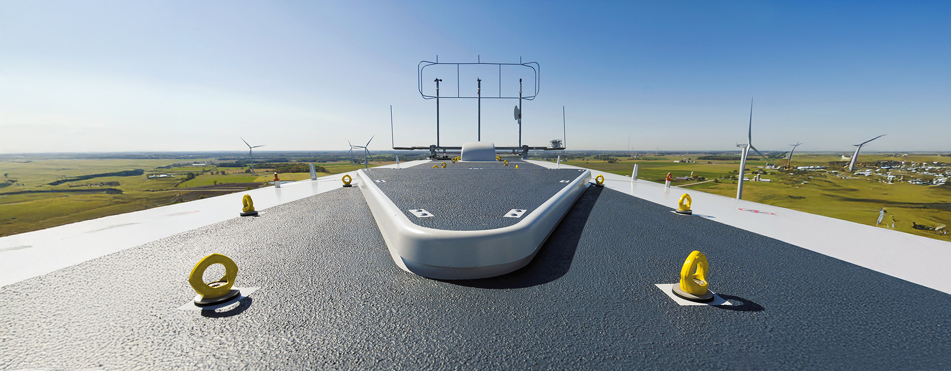 Blick über das Dach einer Windkraftanlage mit mehreren gelben RUD-Personenanschlagpunkten zur Absturzsicherung. Im Hintergrund sind weitere Windräder in einer weiten Landschaft zu sehen, bei klarem Himmel und guter Sicht.