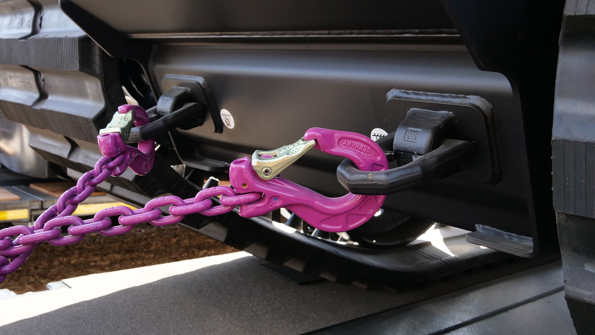 Zwei pinke RUD-Zurrhaken mit Kette sichern ein schweres Fahrzeug auf einem Tieflader; die Haken sind an robusten Befestigungspunkten des Fahrzeugs eingehängt.