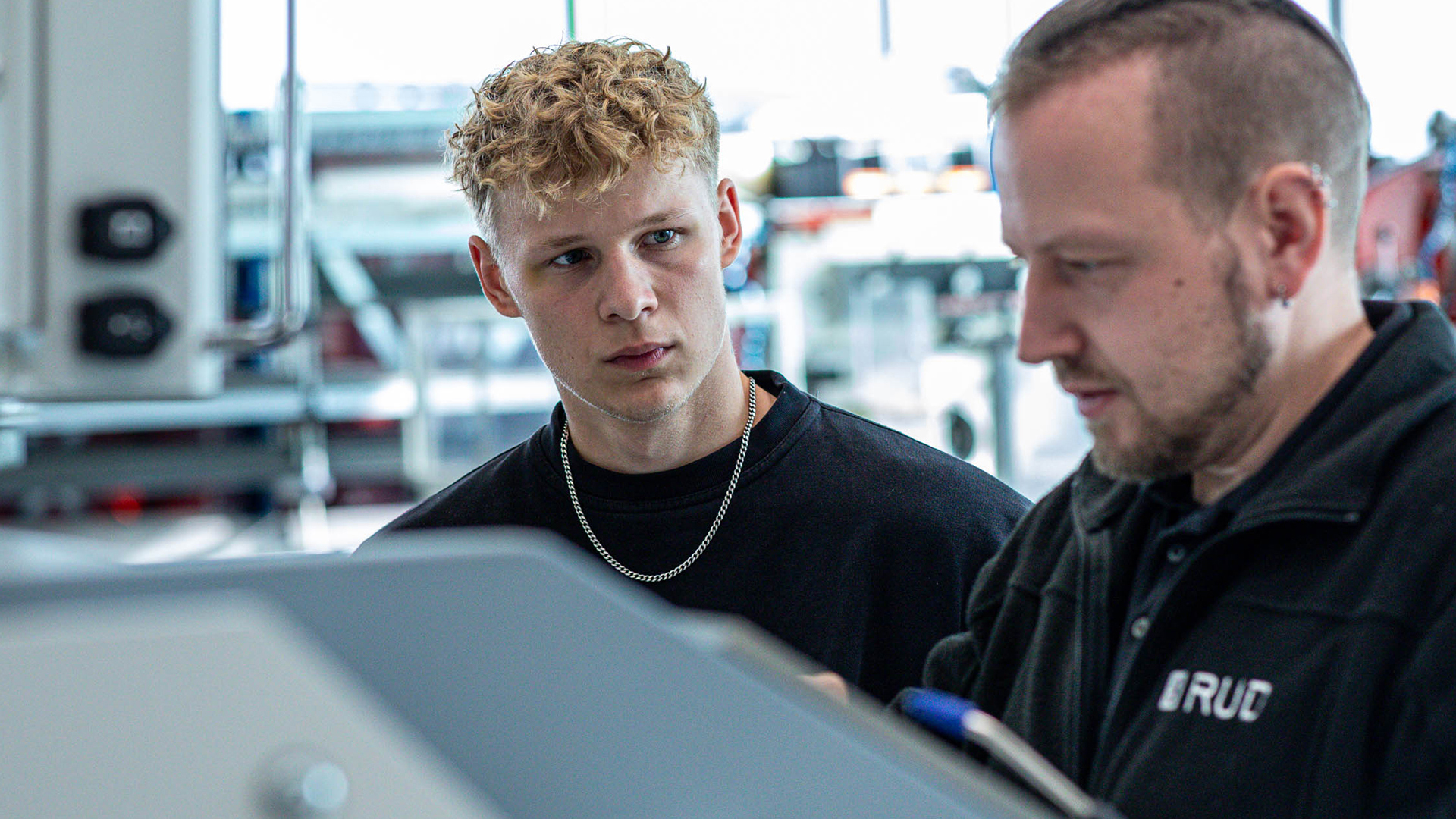 Ein junger Mann beobachtet aufmerksam einen Ausbilder bei der Arbeit an einer Maschine in einer Werkhalle.