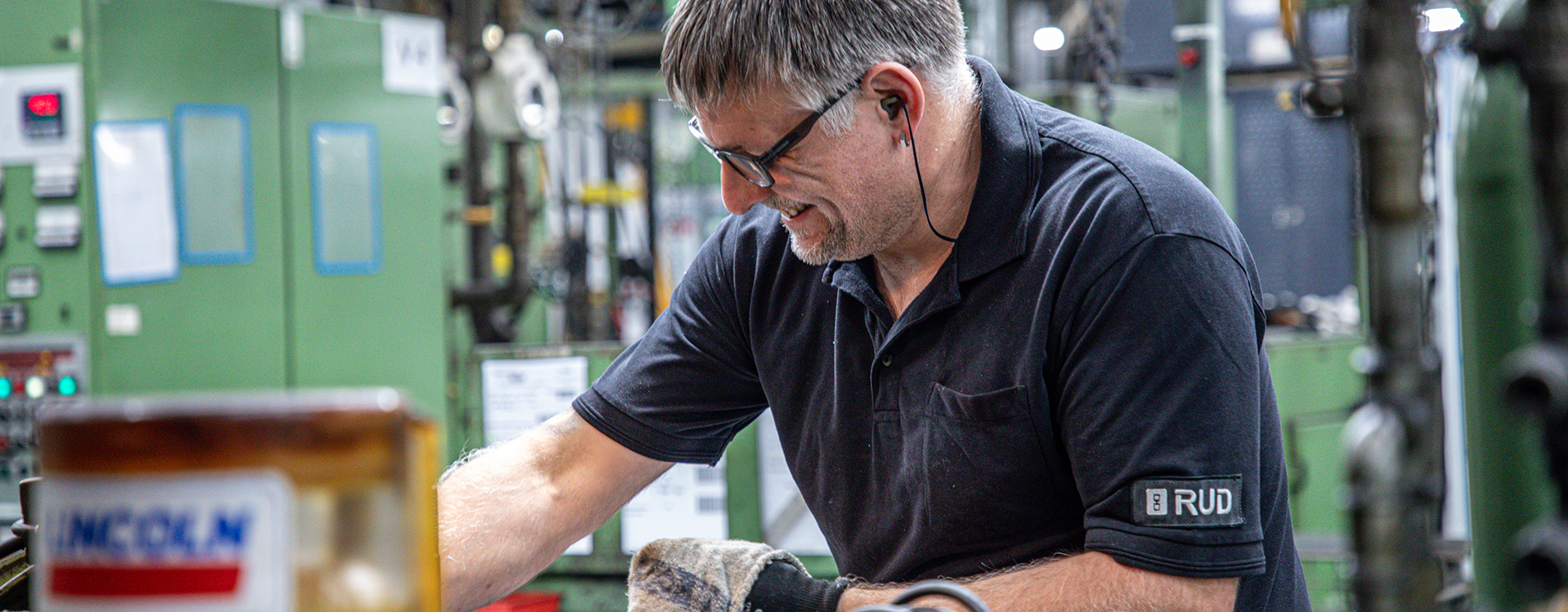 Ein Mitarbeiter in RUD-Arbeitskleidung arbeitet konzentriert an einer Maschine in einer industriellen Produktionshalle.
