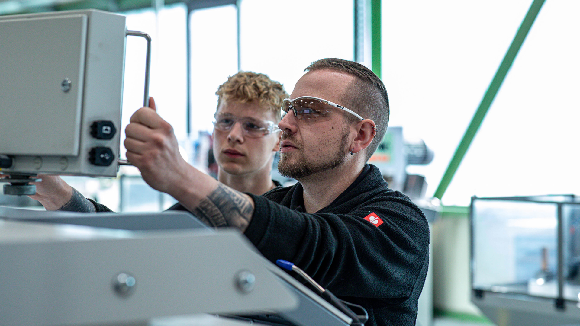 Ein erfahrener RUD-Mitarbeiter erklärt einem jungen Berufseinsteiger einen Arbeitsvorgang an einer Maschine.